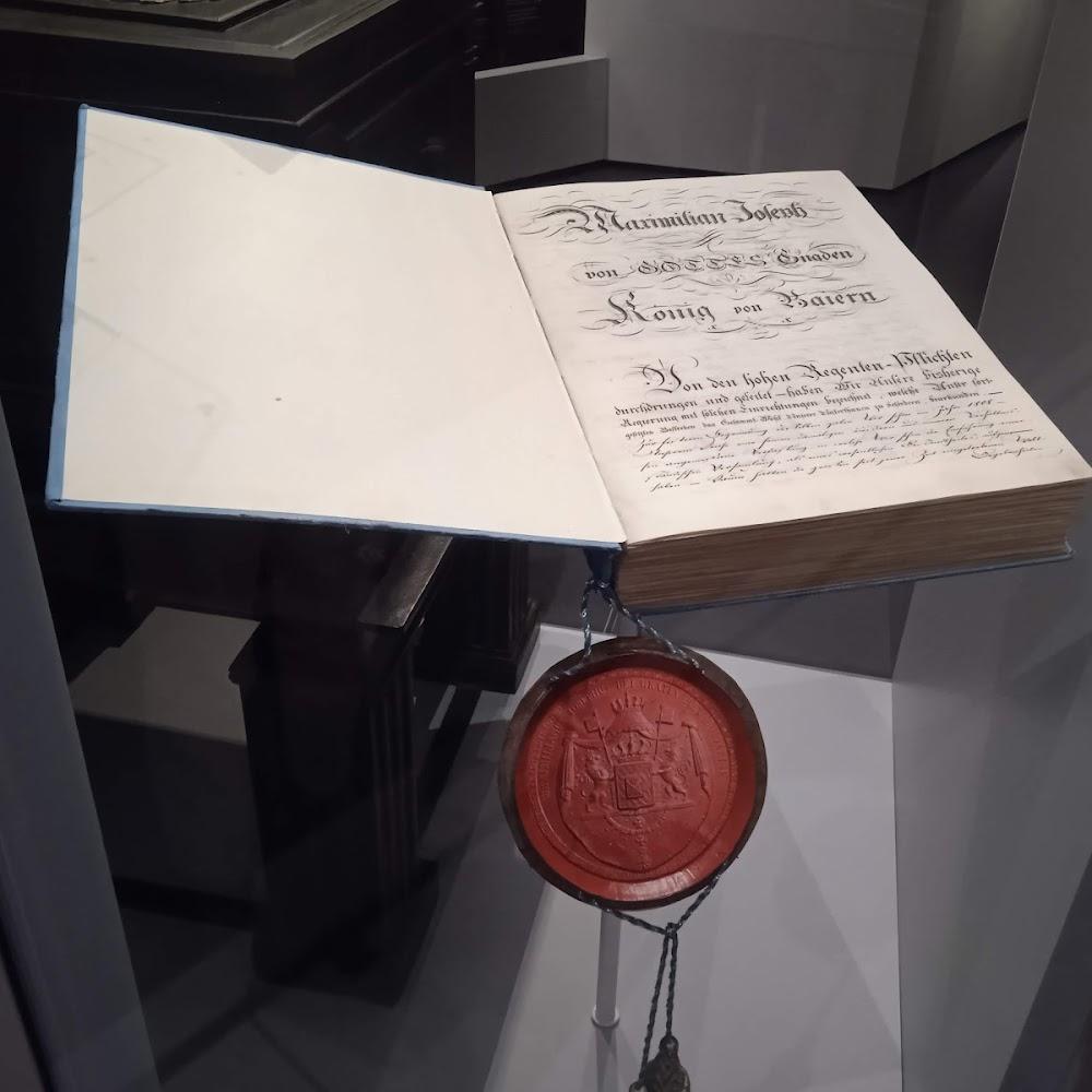 A large, open book with a red wax seal. The book is titled,